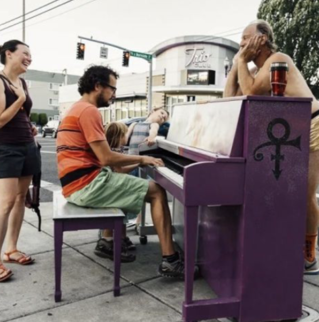 Man playing the piano with people watching and laughing beside him.